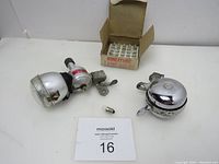 Photo of the vintage bicycle lamp with attached dynamo, the bicycle bell, and the open box of 26 miniature lamp bulbs placed on a white surface with auction number card.
