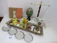 Full lot view showing aluminum coasters, ceramic cow creamer, towel holder, decorative eggs, yellow container, shot glass, mirrored tray, brass bell.
