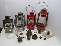 Three vintage oil lamps in red and silver with visible wear and rust, plus loose parts including glass globe, burners, and metal stoppers spread on table.