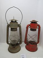 Photo showing two vintage oil lamps side by side: a heavily rusted Beacon lamp with glass and a red Dietz lamp without glass.