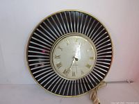 Front view of the clock showing the metal frame with black and silver radial design and the dial with gold Roman numerals and hands, plastic face cover showing a small crack.