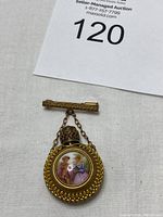 Top view of gold-tone antique perfume brooch with hand-painted Limoges porcelain medallion depicting two women in period dress.
