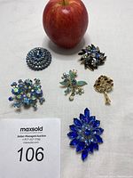 Six brooches arranged on white cloth with apple for scale; multiple styles including blue rhinestones, gold-tone tree, and floral motifs visible.