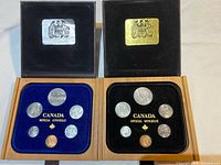 Two coin sets displayed open in wood and leather cases with blue and black velvet interiors, showing complete sets of 7 coins each.