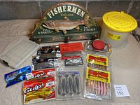 Overview of lot showing various soft bass baits, tackle organizer box, minnow bucket with yellow lid, red bait container, and decorative wall sign with fish relief and hook pegs.