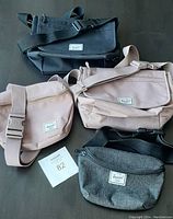 Four Herschel canvas bags laid out: black shoulder bag, dusty blush shoulder bag, large dusty blush waist bag, small dark grey waist bag, all with brand patches clearly visible.