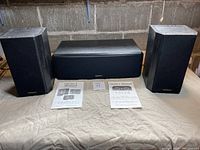 Front view of pair of V52si bookshelf speakers and V52clr centre channel speaker with owners manuals shown on cloth surface.