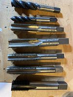 Close-up of 7 machinists taps showing steel construction with spiral and straight flute types, hexagonal shank ends, laid out horizontally.