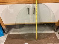 Two identical clear round glass table tops leaning against a wooden cabinet, measuring tape shows approximately 30 inches diameter for each.