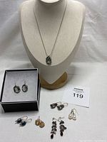 Display of seven pairs of silver metal earrings with various stones and one pendant necklace on bust and box, showing all items in the lot.