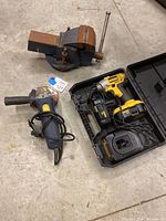 Tools displayed on concrete floor: bench vise, electric angle grinder with power cord, Dewalt impact driver in case with charger and batteries.