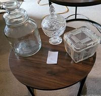 Three glass container items on a round wooden table: large jar, footed candy dish, and square candy dish with lids.