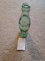 Photo of single green glass Coca-Cola bottle standing upright on carpet with embossed Coca-Cola logo visible.