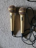 Two gold wired microphones with attached cords and on/off switches lying on carpet.