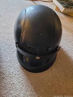 Black motorcycle helmet with visible wear, scuffs and surface dust in indoor setting on carpeted floor