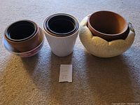 Photo showing three plant holders arranged on a carpet: two nested ceramic pots (brown with black lining) and a white ribbed planter, and a larger decorative textured planter with inset brown pot.