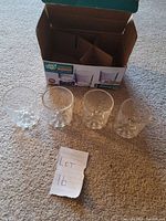 Four clear glass shot glasses displayed in front of open packaging box on carpeted floor with lot number note placed in front.