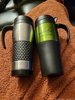 Two travel mugs side by side on a brown fabric surface; one stainless steel with black rubber grip, the other green and black with Peak Rite branding.