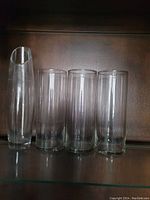 Photo showing four clear glass vases arranged in a row on a dark surface: one tall slender vase and three cylindrical vases.
