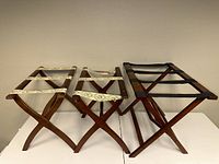 Three wooden luggage valets side by side, showing design and fabric strap patterns.