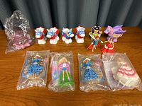 Photo showing all toys on brown surface: four McDonald's Happy Meal dolls sealed in original bags, four white and red cow toys, two small doll figures, and a purple winged pony figure.