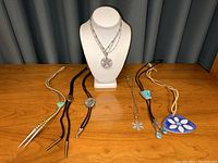 Group image showing all six bolo ties and two layered silver-tone necklaces with flower pendants on white bust form and wooden surface