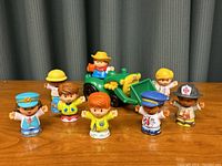 Group of 8 Little People figures arranged with a green and yellow toy tractor in background, captured to show full set and context.