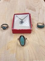 Four silver and turquoise jewelry pieces displayed on a wooden surface. Three rings and one necklace in a red box.