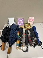 Photo showing nine umbrellas in various colors and styles arranged horizontally, along with two small picture frames and a vintage tin container behind the umbrellas.