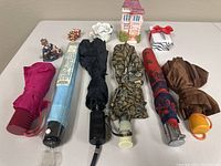 Photo of 6 umbrellas in various colors and patterns arranged in a row on a table, along with several small porcelain trinkets including a miniature house, white flower, and small figurines.