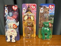 Three Ty Beanie Babies bears (Glory, Britannia, Erin) displayed in original McDonald's blister packaging with themed backdrops.