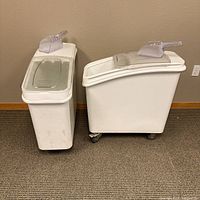 Front view of the pair of white plastic flour bins on wheels with scoops on top, positioned side by side against a wall.
