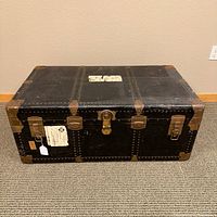 Front view of vintage black steamer trunk with brass corner accents, locks, and rivets, showing wear and patina, with travel labels on top.