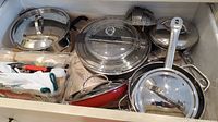 Photo showing assortment of cookware including frying pans, lids, cooking utensils, and a metal steamer insert inside a box.