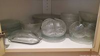 Shelved glass dish set showing stacked dinner and side plates, bowls, cups with saucers, and butter dish.