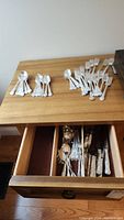 Photo of various silver-plated forks and spoons laid out on a wood table with more flatware in drawer below.