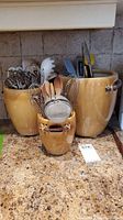 Three ceramic jars on a granite countertop filled with various kitchen utensils including wooden spoons, metal whisks, strainers, and tongs.