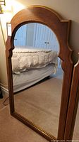 Full frontal photo of a wooden framed hanging mirror showing the arched decorative top and reflective surface, placed in a bedroom setting.