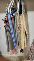 Several pashminas hanging on hangers against a wall, including beige, gray, blue, and others showing different textures and patterns with some fringes and tassels.