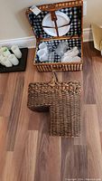 Top view of open wicker picnic basket showing black and white checkered lining, plates, cups and glasses inside, and closed decorative wicker basket next to it on floor.