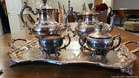 Full view of the 5 piece silver plated tea service set placed on a wooden surface including tray, teapot, coffee pot, creamer, and sugar bowl.
