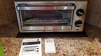 Full front view of the toaster oven on a kitchen counter along with the manual and an auction bid card.
