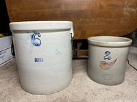 Two stoneware crocks side by side on carpeted floor, 6 gallon Ruckel's on left and 2 gallon Red Wing on right
