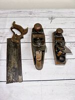 Three antique woodworking hand tools on white wooden floor: hand saw and two different sized wood planes.