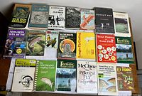 Photo shows 20 books spread out on a surface including fishing guides, techniques, an alchemy book, and a signed Rocket Men first edition.