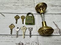 Seven assorted vintage keys, one metal padlock marked REESE, and ornate brass doorknob on a wood background