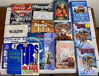 Overview photo showing all books and magazines flat on table with titles visible, including George R.R. Martin's 'A Storm of Swords',' Robert Jordan novels, and various collectibles and hobby magazines.
