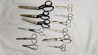 Full set of 10 various scissors on white fabric background showing variety in size, handle styles and blade shapes