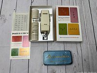 Full contents including Singer Monogrammer device, alphabet bands, instruction manual, and blue metal storage box all arranged with original colorful packaging box and white foam insert