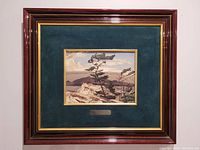Frontal photo of the framed print showing the entire item including dark wood frame with gold trim and dark green matte surrounding the print. Small metal plaque below the image visible.